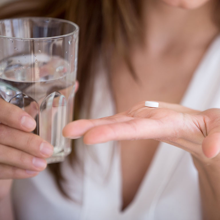 Frau hält Tablette in der einen Hand und möchte sie mit einem Glas Wasser einnehmen, um ihre Blasenentzündung zu behandeln.